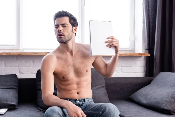 Sexy shirtless man with closed eyes suffering from heat while waving with notepad as fan at home — Stock Photo