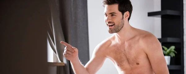 Panoramic shot of handsome smiling shirtless man in jeans pointing at home — Stock Photo