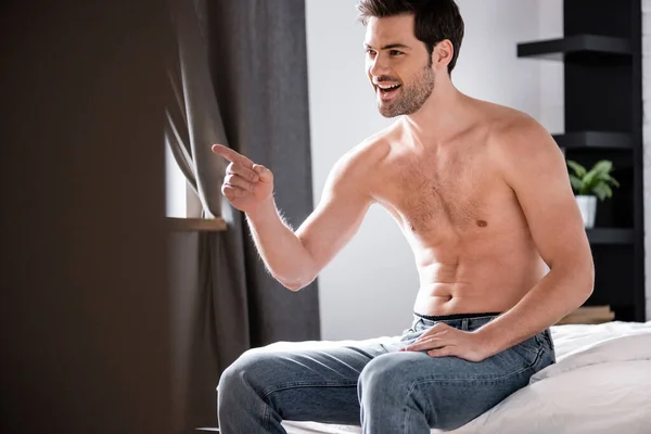 Bel homme torse nu et souriant en jeans pointant du doigt tout en étant assis sur le lit — Photo de stock
