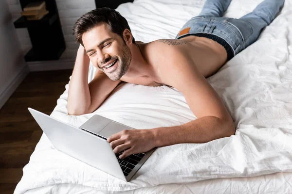 Красивый мужчина без рубашки смеется и использует ноутбук на кровати — стоковое фото