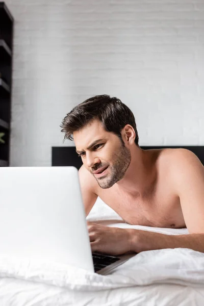 Dissatisfied shirtless male freelancer working on laptop in bed — Stock Photo