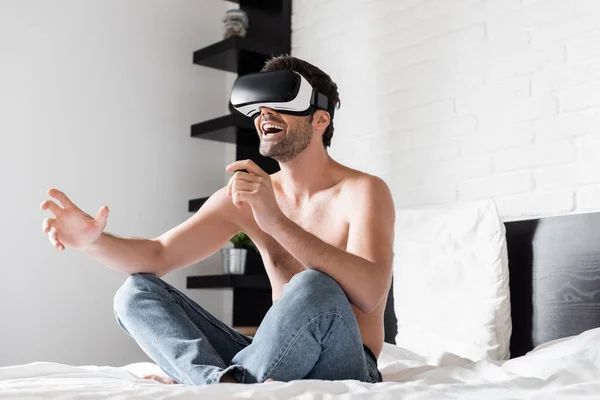 Возбужденный мужчина без рубашки жесты при использовании виртуальной реальности гарнитуры на кровати — стоковое фото