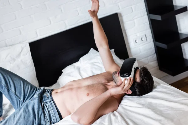 Homem sem camisa gesticulando enquanto deitado e usando fone de ouvido realidade virtual na cama — Fotografia de Stock