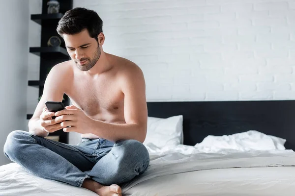 Позитивный мужчина без рубашки с помощью смартфона в спальне дома — стоковое фото