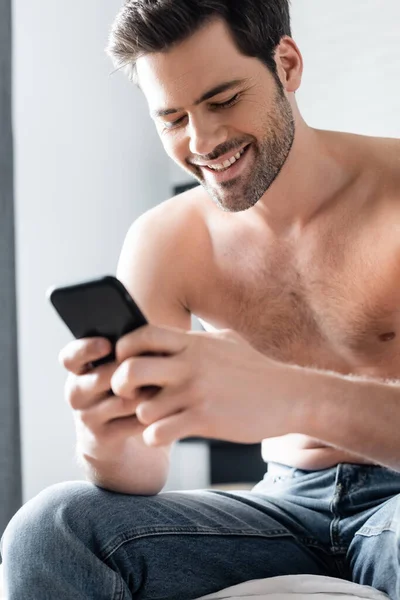 Улыбающийся сексуальный мужчина без рубашки с помощью смартфона на кровати дома — стоковое фото