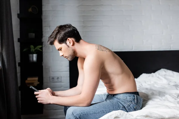 Молодой мужчина без рубашки с помощью смартфона на кровати — стоковое фото