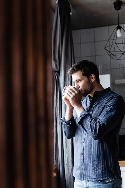 Schöner Mann trinkt Kaffee, während er zu Hause durch das Fenster auf Quarantäne schaut — Stockfoto