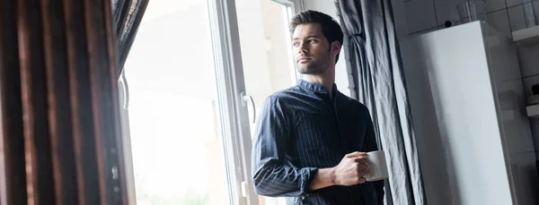 Homem bonito segurando xícara de café e de pé perto da janela em casa em quarentena, conceito horizontal — Fotografia de Stock