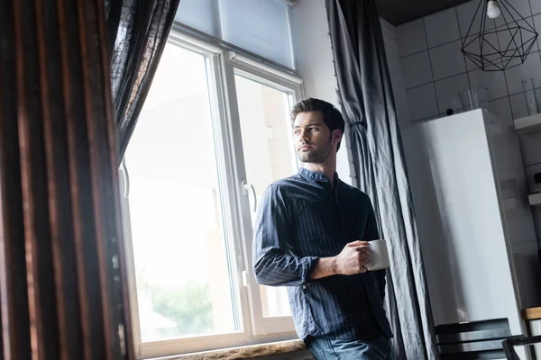 Guapo pensativo hombre sosteniendo taza de café y de pie cerca de la ventana en casa en auto aislamiento - foto de stock