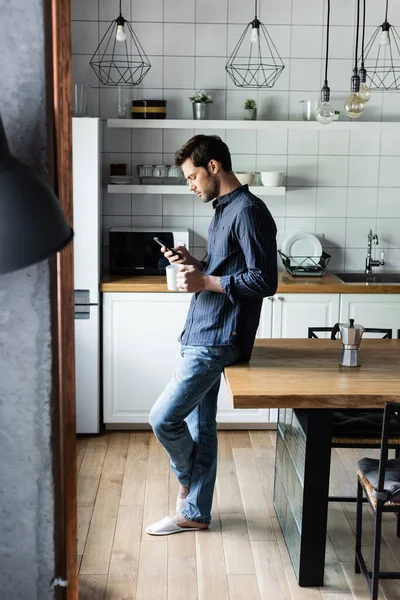 Homem bonito segurando xícara de café enquanto usa smartphone na cozinha durante a quarentena — Fotografia de Stock