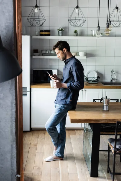 Bell'uomo che tiene in mano una tazza di caffè mentre usa lo smartphone in cucina durante la quarantena — Foto stock