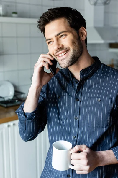 Cheerful handsome man holding cup of coffee while talking on phone on kitchen during quarantine — Stock Photo
