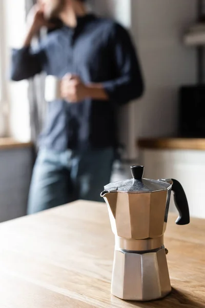 Vista recortada del hombre sosteniendo la taza de café y hablando por teléfono celular en la cocina con cafetera durante la cuarentena, enfoque selectivo — Stock Photo