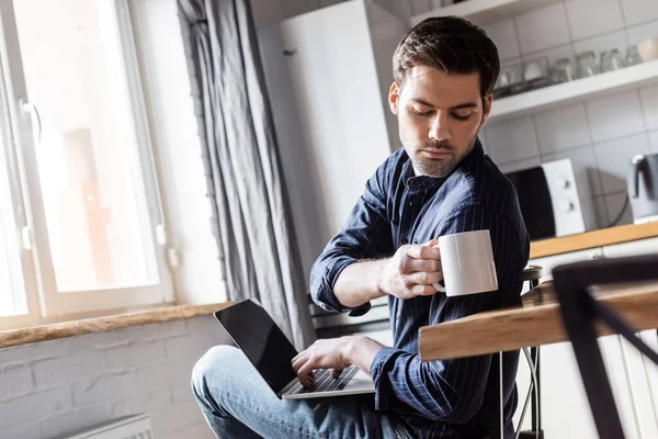 Красивый мужчина держит чашку кофе, используя ноутбук на кухне во время карантина — стоковое фото