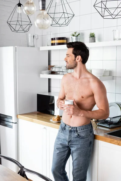 Sonriente hombre sin camisa sosteniendo taza de café en la cocina por la mañana - foto de stock