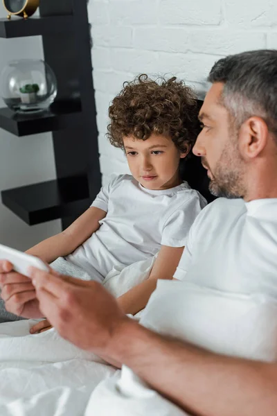 Selective focus of handsome father holding smartphone and looking at curly son — Stock Photo