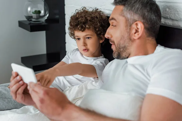 Selektiver Fokus des niedlichen Jungen, der mit dem Finger auf sein Smartphone in der Nähe des Vaters zeigt — Stockfoto