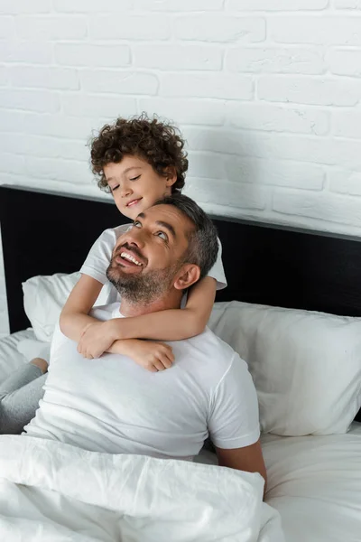 Niño rizado abrazando feliz padre en el dormitorio - foto de stock