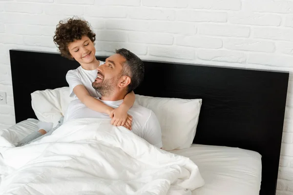 Niño rizado abrazando alegre padre en el dormitorio - foto de stock