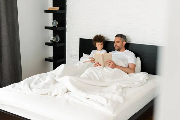 Selective focus of happy father and curly son reading book in bedroom — Stock Photo