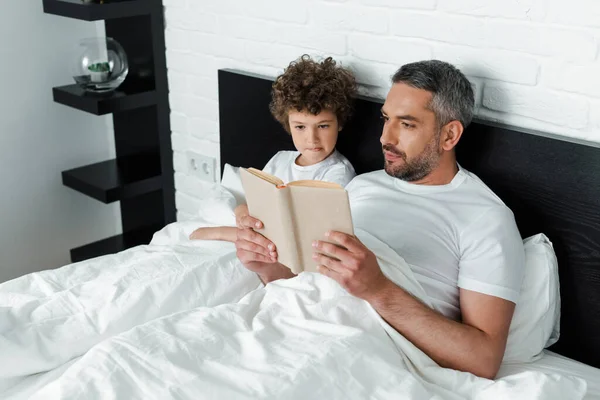 Bärtiger Vater liest Buch neben lockigem Sohn im Schlafzimmer — Stockfoto