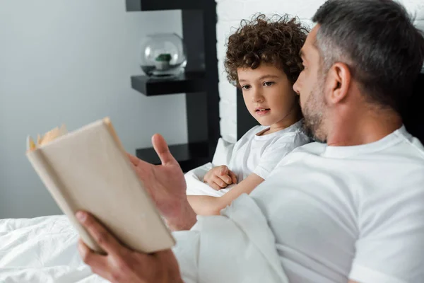 Selective focus of curly boy looking at book near father in bedroom — Stock Photo
