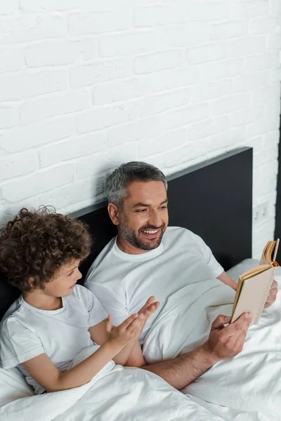 Cheerful father reading book near curly son in bedroom — Stock Photo