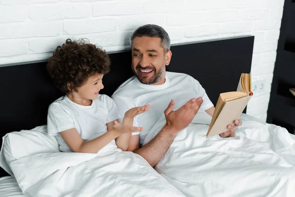 Кудрявый мальчик жестикулирует рядом с счастливым отцом, держа книгу в спальне — стоковое фото