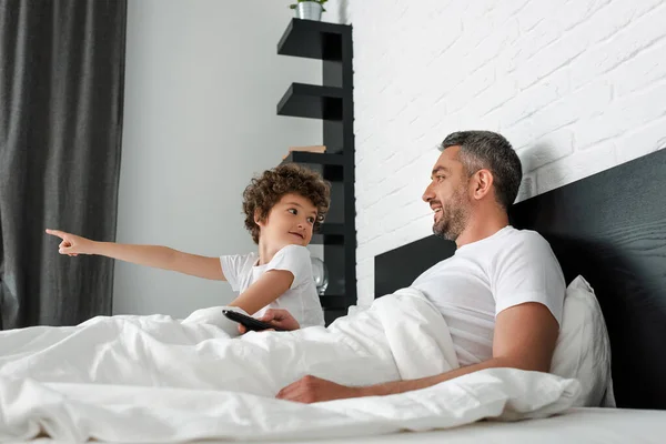 Enfoque selectivo de chico lindo señalando con el dedo y mirando a padre con mando a distancia - foto de stock