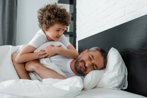 Menino encaracolado olhando para o pai barbudo com os olhos fechados deitado na cama — Fotografia de Stock