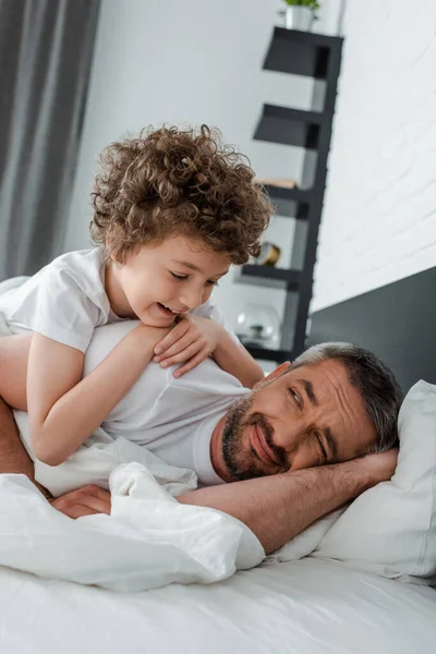 Niño rizado mirando al padre barbudo acostado en la cama - foto de stock