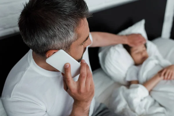 Enfoque selectivo de padre tocando enfermo hijo y hablando en el teléfono inteligente en el dormitorio - foto de stock