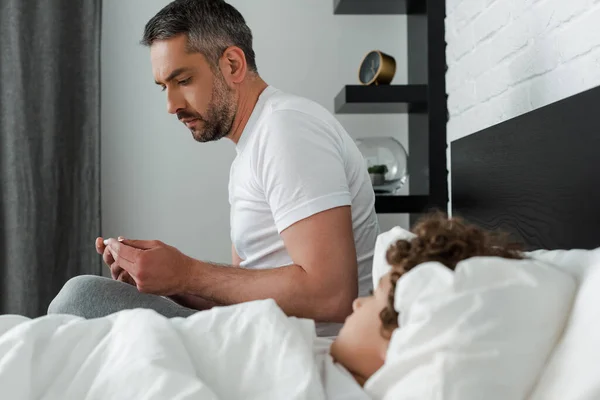 Selective focus of worried father holding digital thermometer near sick son in bed — Stock Photo