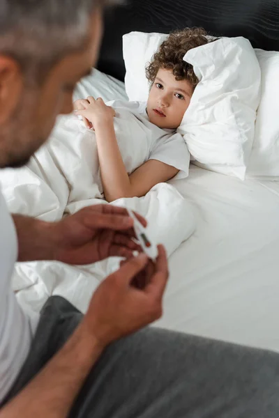 Enfoque selectivo de niño enfermo mirando a padre con termómetro digital - foto de stock