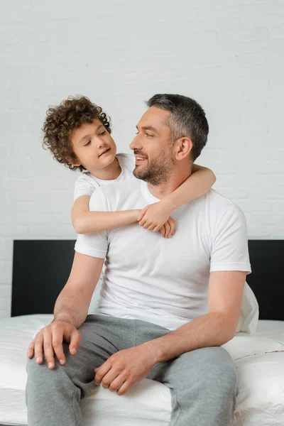 Кудрявый сын обнимает счастливого бородатого отца и смотрит друг на друга в спальне — стоковое фото
