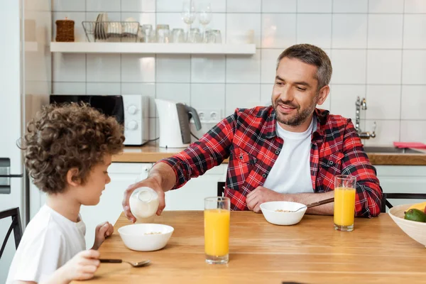 Messa a fuoco selettiva di felice padre bottiglia con latte vicino figlio carino, bicchieri di succo d'arancia e ciotola con fiocchi di mais — Foto stock