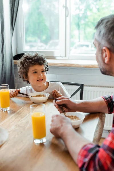 Enfoque selectivo de niño alegre mirando a padre cerca de cuencos con copos de maíz y vasos de jugo de naranja - foto de stock