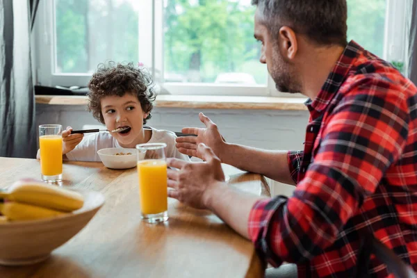 Foco seletivo do menino encaracolado comendo flocos de milho e olhando para o pai perto de tigela e copos de suco de laranja — Fotografia de Stock