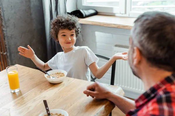 Foyer sélectif de garçon bouclé gesticulant et regardant le père près des bols avec des flocons de maïs et un verre de jus d'orange — Photo de stock