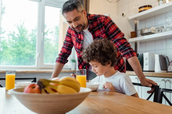 Selektiver Fokus des gutaussehenden Vaters, der neben dem lockigen Sohn steht und auf dem Tisch frühstückt — Stockfoto