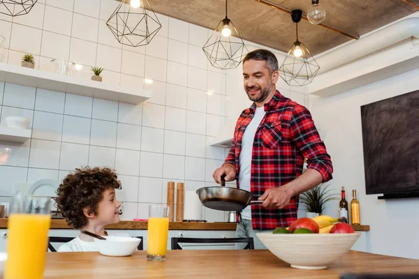 Избирательный фокус счастливого мальчика, смотрящего на веселого отца, держащего сковородку на кухне — стоковое фото
