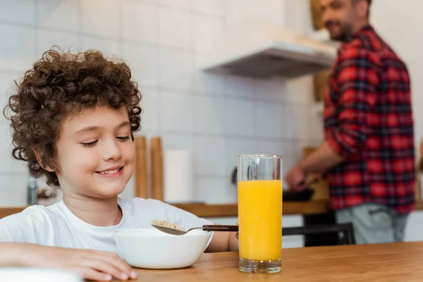 Fuoco selettivo di ragazzo allegro e riccio che tiene il cucchiaio vicino a boccia e vetro di succo d'arancia in cucina — Foto stock