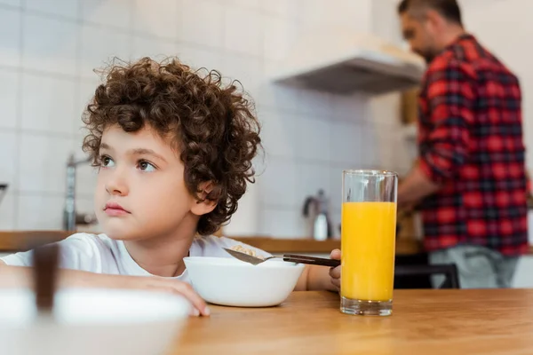 Foyer sélectif de garçon coûteux et bouclé regardant loin près du petit déjeuner et le père dans la cuisine — Photo de stock