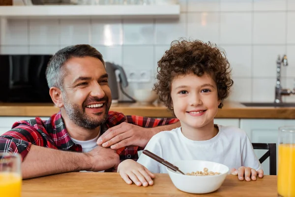 Foco seletivo de pai alegre olhando para o filho sorridente e encaracolado perto do café da manhã na mesa — Fotografia de Stock