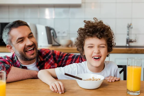 Foco seletivo de pai alegre olhando para o filho bonito e encaracolado perto do café da manhã na mesa — Fotografia de Stock