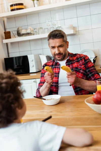 Foco seletivo do pai segurando bananas frescas enquanto brincava com o filho encaracolado perto do café da manhã na mesa — Fotografia de Stock