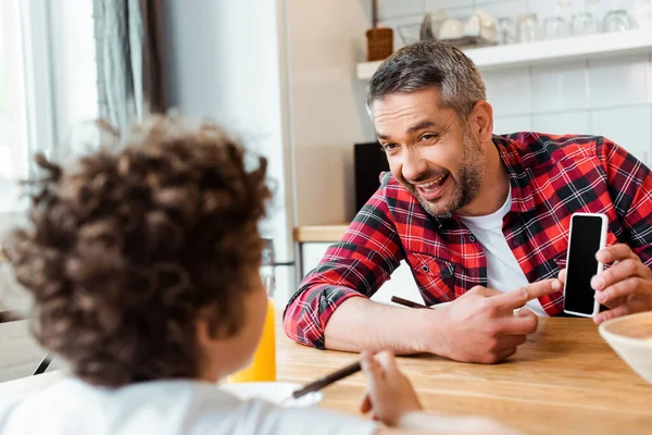 Messa a fuoco selettiva del padre felice che punta con il dito allo smartphone con schermo bianco vicino al figlio riccio — Foto stock