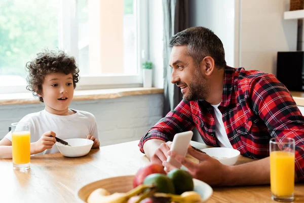 Избирательный фокус счастливого отца, держащего смартфон рядом удивленный сын на кухне — стоковое фото