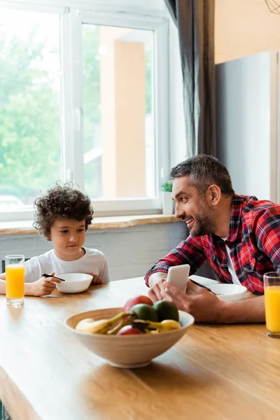 Избирательный фокус счастливого отца, держащего смартфон рядом с милым сыном на кухне — стоковое фото