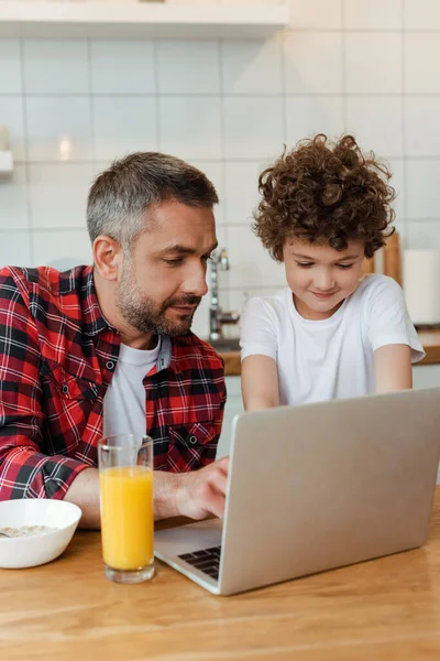 Enfoque selectivo del padre freelancer guapo y el hijo rizado utilizando el ordenador portátil en la cocina - foto de stock